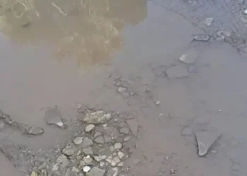 Pozza d'acqua torbida su asfalto dissestato, pietre e detriti a terra, recinzione in rete metallica sullo sfondo