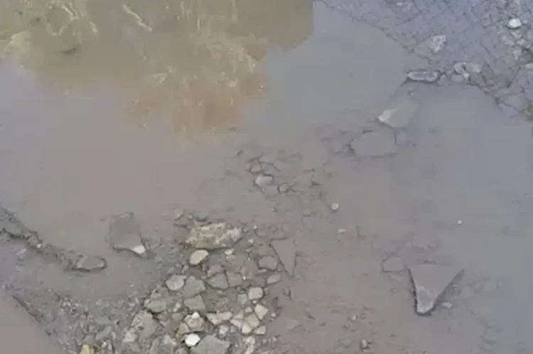 Pozza d'acqua torbida su asfalto dissestato, pietre e detriti a terra, recinzione in rete metallica sullo sfondo