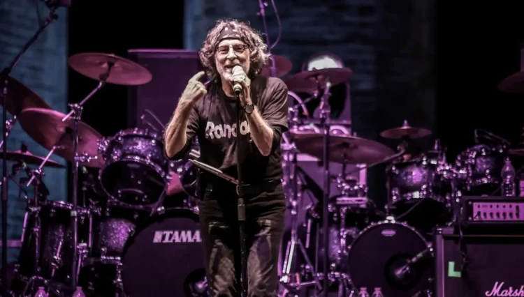 Un cantante con microfono sul palco, luci viola, batterie Tama sullo sfondo durante un concerto rock