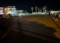 Piazzetta del porto turistico di Marina di Ragusa di notte, con gran parte della pavimentazione in penombra e palme illuminate sullo sfondo.