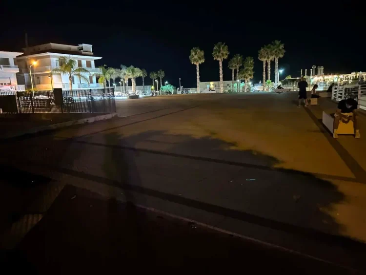 Piazzetta del porto turistico di Marina di Ragusa di notte, con gran parte della pavimentazione in penombra e palme illuminate sullo sfondo.