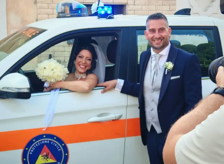 Sposi in posa accanto a un mezzo della Protezione Civile: la sposa al finestrino con bouquet, lo sposo in abito blu.