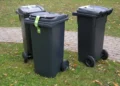 Tre bidoni carrellati grigio scuro con nastro di sigillo verde sul coperchio, su un prato accanto a un vialetto