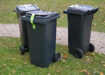 Tre bidoni carrellati grigio scuro con nastro di sigillo verde sul coperchio, su un prato accanto a un vialetto