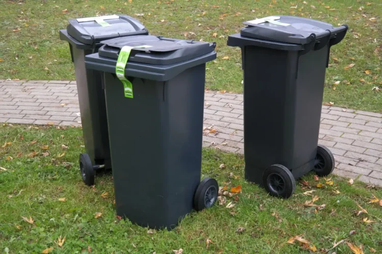 Tre bidoni carrellati grigio scuro con nastro di sigillo verde sul coperchio, su un prato accanto a un vialetto