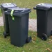 Tre bidoni carrellati grigio scuro con nastro di sigillo verde sul coperchio, su un prato accanto a un vialetto