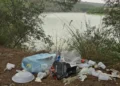 Sacchetti e contenitori di plastica abbandonati tra pini e vegetazione, sullo sfondo lo specchio d'acqua della diga
