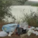 Sacchetti e contenitori di plastica abbandonati tra pini e vegetazione, sullo sfondo lo specchio d'acqua della diga