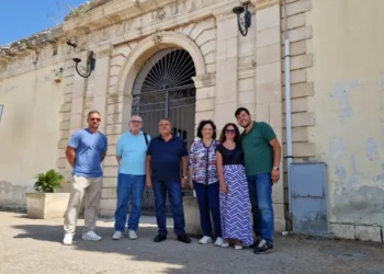 Sei persone in posa davanti al portale d'ingresso del cimitero comunale di Comiso, con intonaco ammalorato visibile sulla facciata.