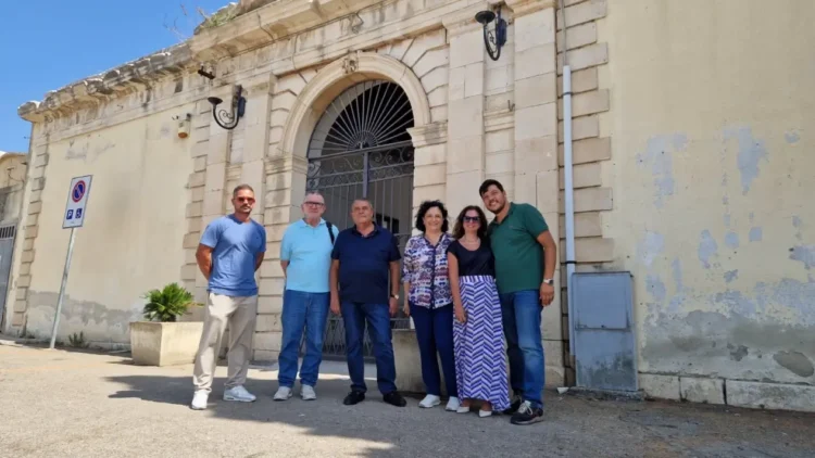 Sei persone in posa davanti al portale d'ingresso del cimitero comunale di Comiso, con intonaco ammalorato visibile sulla facciata.