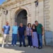 Sei persone in posa davanti al portale d'ingresso del cimitero comunale di Comiso, con intonaco ammalorato visibile sulla facciata.