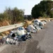 Cumuli di rifiuti abbandonati lungo il guardrail di una strada, con sterpaglie e alberi sulla carreggiata