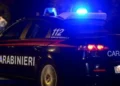 Auto dei Carabinieri con lampeggianti blu accesi in una strada buia, in secondo piano due persone in piedi sul marciapiede
