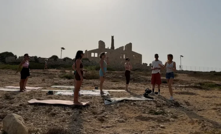 Gruppo di persone su tappetini da yoga davanti alle rovine della Fornace di Punta Pisciotto a Sampieri, Scicli