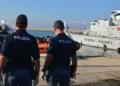 Due agenti di Polizia di spalle osservano l'arrivo di migranti a bordo di un gommone arancione nel porto, accanto a una nave della Guardia di Finanza