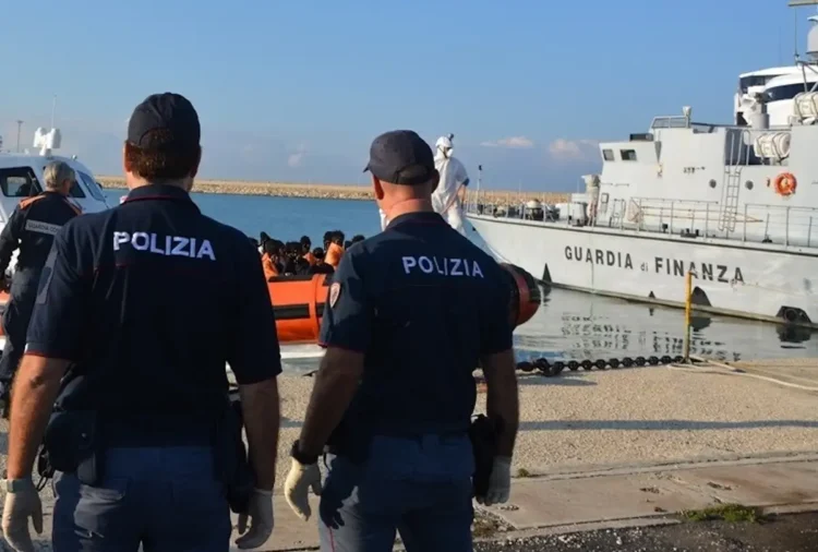 Due agenti di Polizia di spalle osservano l'arrivo di migranti a bordo di un gommone arancione nel porto, accanto a una nave della Guardia di Finanza