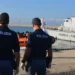 Due agenti di Polizia di spalle osservano l'arrivo di migranti a bordo di un gommone arancione nel porto, accanto a una nave della Guardia di Finanza