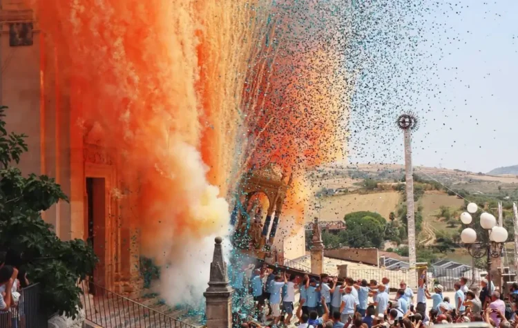 Portatori in maglietta azzurra sorreggono il simulacro della Madonna della Neve tra nuvole di fumo arancione e coriandoli colorati davanti alla basilica di Giarratana