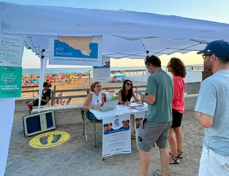 Gazebo dell'ASP di Ragusa su un lungomare affollato: due operatrici allo sportello accolgono cittadini per lo screening oncologico