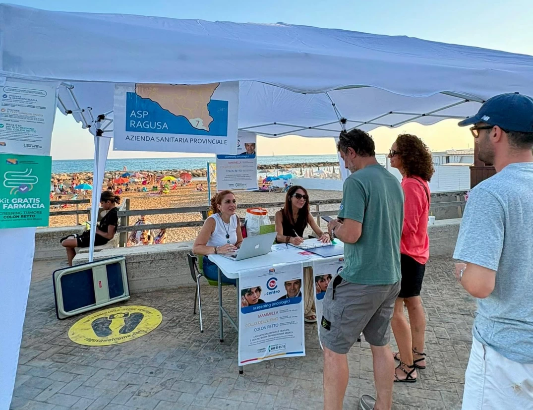 Gazebo dell'ASP di Ragusa su un lungomare affollato: due operatrici allo sportello accolgono cittadini per lo screening oncologico