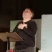 Amedeo Fusco recita in piedi davanti a un leggio durante uno spettacolo teatrale.