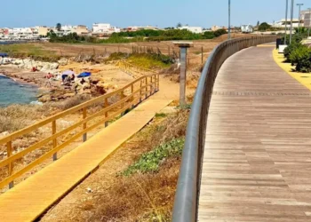 Passerella pedonale in legno a Punta di Mola, Marina di Ragusa, con accesso alla spiaggia rocciosa e il lungomare sullo sfondo