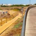 Passerella pedonale in legno a Punta di Mola, Marina di Ragusa, con accesso alla spiaggia rocciosa e il lungomare sullo sfondo