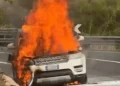 Suv in fiamme sul bordo della strada, alte fiamme dal cofano anteriore e fumo nero, guardrail sullo sfondo.