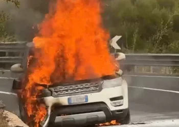 Suv in fiamme sul bordo della strada, alte fiamme dal cofano anteriore e fumo nero, guardrail sullo sfondo.