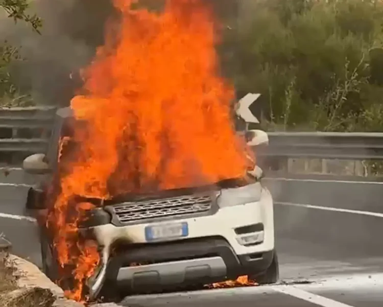 Suv in fiamme sul bordo della strada, alte fiamme dal cofano anteriore e fumo nero, guardrail sullo sfondo.