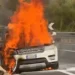 Suv in fiamme sul bordo della strada, alte fiamme dal cofano anteriore e fumo nero, guardrail sullo sfondo.