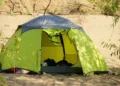 Tenda da campeggio verde montata sulla sabbia di una spiaggia, con vegetazione intorno e bagagli visibili all'interno