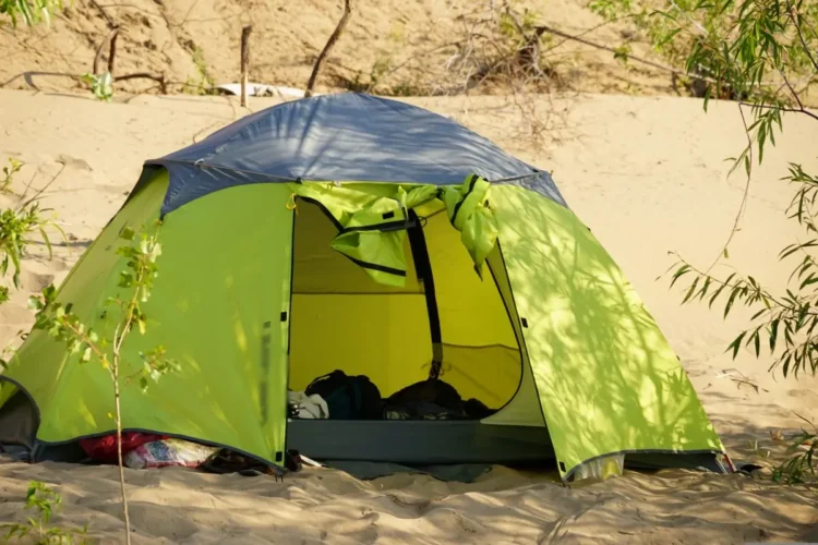 Tenda da campeggio verde montata sulla sabbia di una spiaggia, con vegetazione intorno e bagagli visibili all'interno