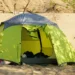 Tenda da campeggio verde montata sulla sabbia di una spiaggia, con vegetazione intorno e bagagli visibili all'interno