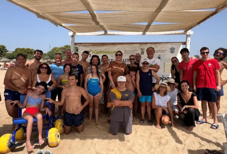 Ragazzi, famiglie e volontari in spiaggia davanti al gazebo della Base Nautica, uno su sedia a rotelle da mare
