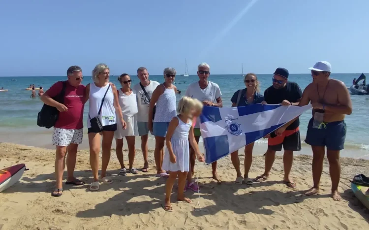 Gruppo di soci in spiaggia con il gonfalone della Lega Navale Italiana, barche a vela sullo sfondo a Marina di Ragusa