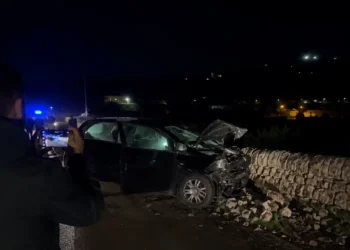 Auto incidentata di notte contro un muro a secco, un uomo la riprende con lo smartphone, luci blu dei soccorsi sullo sfondo
