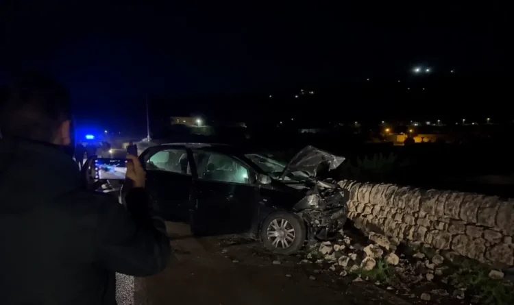 Auto incidentata di notte contro un muro a secco, un uomo la riprende con lo smartphone, luci blu dei soccorsi sullo sfondo