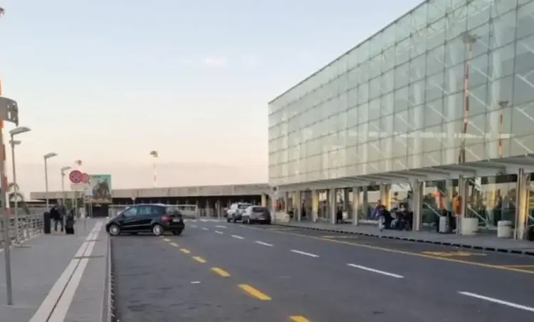 Ingresso esterno dell'aeroporto di Catania Fontanarossa con terminal in vetro, auto in sosta e passeggeri sul marciapiede.