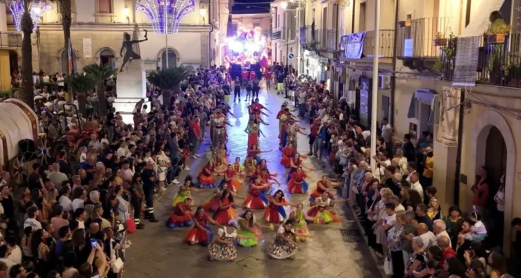 Ballerini in costume colorato si esibiscono tra la folla in una via illuminata di Chiaramonte Gulfi durante Zuppà.