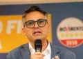 Sergio Firrincieli parla al microfono durante un evento pubblico, con manifesti elettorali sullo sfondo