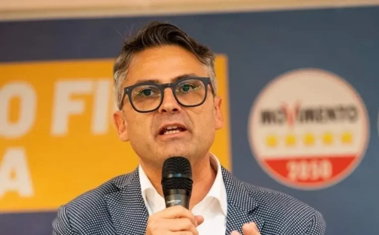 Sergio Firrincieli parla al microfono durante un evento pubblico, con manifesti elettorali sullo sfondo