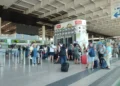 Passeggeri con trolley in fila all'aeroporto di Catania davanti al totem degli orari dei voli, vicino ai banchi check-in 16-20.