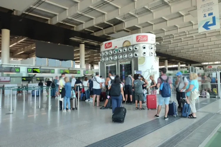 Passeggeri con trolley in fila all'aeroporto di Catania davanti al totem degli orari dei voli, vicino ai banchi check-in 16-20.