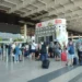 Passeggeri con trolley in fila all'aeroporto di Catania davanti al totem degli orari dei voli, vicino ai banchi check-in 16-20.