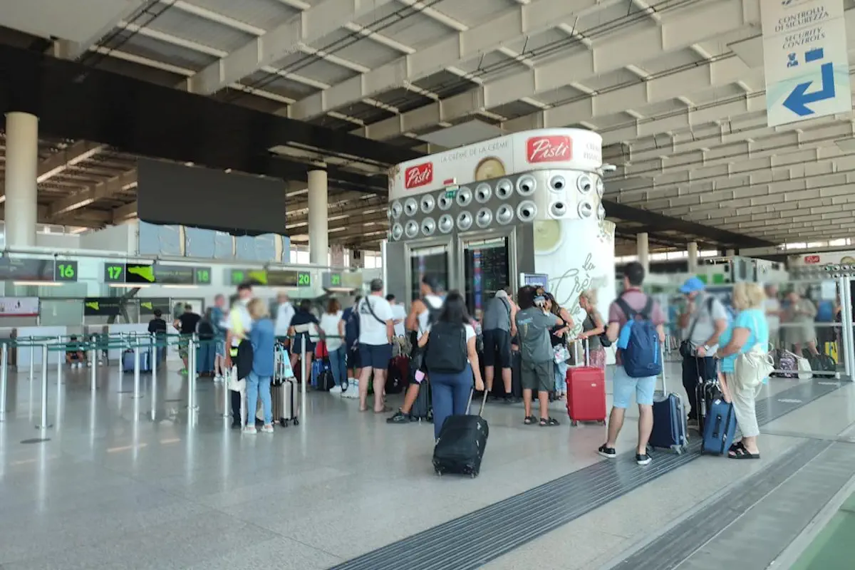 Passeggeri con trolley in fila all'aeroporto di Catania davanti al totem degli orari dei voli, vicino ai banchi check-in 16-20.