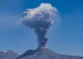 Colonna di cenere grigia che sale da un vulcano sotto un cielo blu, con un altopiano pietroso in primo piano