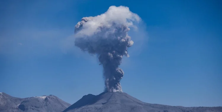 Colonna di cenere grigia che sale da un vulcano sotto un cielo blu, con un altopiano pietroso in primo piano