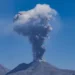 Colonna di cenere grigia che sale da un vulcano sotto un cielo blu, con un altopiano pietroso in primo piano