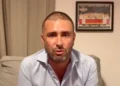 Alessandro Di Battista seduto su un divano grigio, camicia azzurra, con una cicatrice visibile sul capo rasato